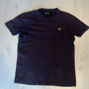 Mörkblå t-shirt från Lyle & Scott - Säljer en mörkblå t-shirt från Lyle & Scott med korta ärmar och ett broderat gult emblem på bröstet. Perfekt för en avslappnad stil. T-shirten är i bomull och har en klassisk passform.