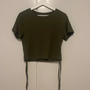 Olivgrön ribbad topp - Snygg olivgrön ribbad topp med korta ärmar och knytband i sidorna. Perfekt för en avslappnad stil med en modern touch. Toppen är croppad och har en bekväm passform.