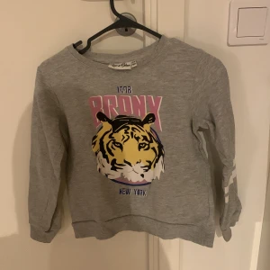 Grå sweatshirt med tigertryck från Red & Blue - Säljer en cool grå sweatshirt från Red & Blue med ett stort tigertryck och texten 'Bronx New York' på framsidan. Tröjan har långa ärmar med vita ränder på ena ärmen. Perfekt för en avslappnad stil!