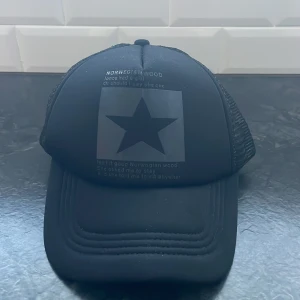 Svart keps med stjärna och text - Snygg svart keps med en stor stjärna och texten 'Norwegian Wood' på framsidan. Kepsen har ett nätmaterial på baksidan för bättre ventilation och en justerbar rem för perfekt passform.