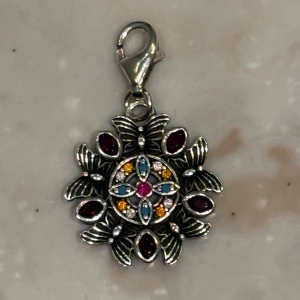 Charm från Thomas Sabo - Charm i silver med färgglada stenar i lila, blått och gult. Med fjärilar i en cirkel med massa stenar runtom. Passar bra till ett armband eller halsband! Inte fast pris! Halsbandet säljs inte längre. Halsbandet är i bra skick och har rengjorts i butik nyligen då hängstycket byttes ut. 