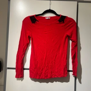 Röd långärmad topp med spetsdetaljer - Snygg röd långärmad topp med svarta spetsdetaljer vid axlarna. Toppen har en rund halsringning och är tillverkad i ett mjukt material.