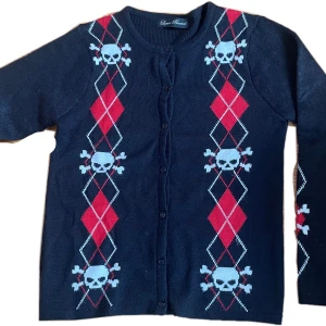 Svart kofta med dödskallemönster från Liquor Brand - Snygg vintage stickad svart kofta från Liquor Brand med ett coolt dödskallemönster i vitt och rött. Koftan har långa ärmar  knappar framtill. Perfekt för dig som vill ha en unik stil! Skulle säga runt storlek s. Köpt runt 2005