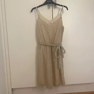 Champagnefärgad klänning - Elegant champagnefärgad klänning med smala axelband och ett knytbälte i midjan. Klänningen har en subtil glittrig yta och är perfekt för speciella tillfällen. Den är ärmlös och har en lätt och luftig känsla.