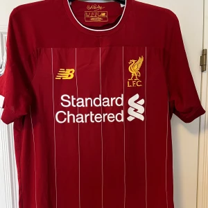 Röd Liverpool fotbollströja från New Balance - Säljer en röd Liverpool fotbollströja från New Balance med vita detaljer och klubbens emblem på bröstet. Tröjan har korta ärmar och sponsorlogga på framsidan. På baksidan står 'VIRGIL' och nummer 4. Perfekt för fans av laget!