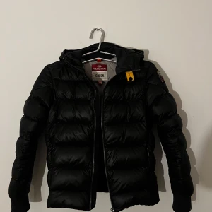 Svart dunjacka från Parajumpers - Säljer en snygg svart dunjacka från Parajumpers med huva och dragkedja. Jackan har en klassisk pufferdesign och en liten logotyp på ärmen. Perfekt för både vinter vår och höst, storleken är y-m som är en barn modell, Hör av er för fler bilder, den är i väldigt fint skick o har inga defekter alls