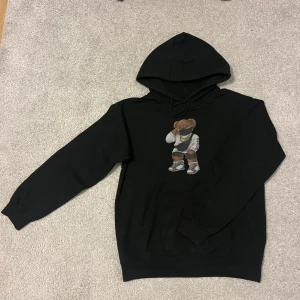 Svart hoodie från Fashionbear - Säljer en svart hoodie från Fashionbear med ett coolt tryck av en björn i streetwear-stil på framsidan. Hoodien har en klassisk passform med långa ärmar och en justerbar huva. Perfekt för en avslappnad look. Köpt för ungefär 600 kr
