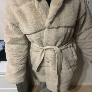 Beige teddyjacka med bälte - Mysig beige teddyjacka med bälte i midjan. Jackan har en fluffig och varm design, perfekt för kyliga dagar. Den har långa ärmar och en huva för extra komfort. Perfekt för att hålla stilen och värmen i vinter.