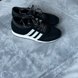 Svarta sneakers från Adidas - Snygga svarta sneakers från Adidas med vita ränder och snörning. Perfekta för en avslappnad stil. Skorna har en vit sula som ger en fin kontrast mot det svarta tyget.