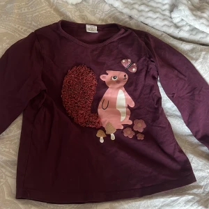 Vinröd långärmad tröja från Lindex Kids - Söt vinröd långärmad tröja från Lindex Kids med ett gulligt ekorrmotiv på framsidan. Ekorrens svans är dekorerad med volangdetaljer och det finns små svampar i olika färger. Perfekt för höst och vinter!