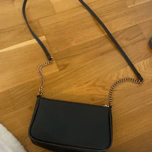 Svart axelväska med kedjedetaljer - Snygg svart axelväska med guldfärgade kedjedetaljer och dragkedja upptill. Perfekt för att bära det viktigaste med stil. Väskan har en smidig axelrem och en kompakt design som passar till många olika outfits.