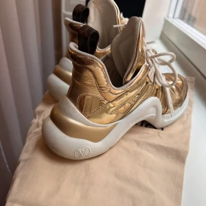 Guldiga sneakers med snörning - Snygga guldiga LV Archlight sneakers med vit sula och snörning. Skorna har en unik design med hög sula och detaljer i guld. Perfekta för att ge en glittrig touch till din outfit.
