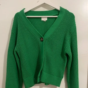 Grön stickad kofta från H&M - Säljer en snygg grön stickad kofta från H&M. Koftan har en djup v-ringning och knäpps med stora knappar framtill. Perfekt för att hålla sig varm och stilren under kyligare dagar.