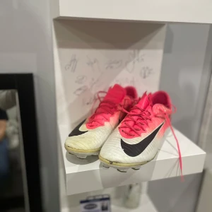 Nike mercurial  - Snygga fotbollsskor från Nike i vitt med rosa detaljer och svart swoosh. Skorna har snörning och är perfekta för fotbollsplanen. De har en klassisk design med dobbar för bra grepp.