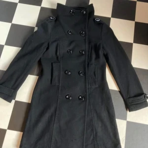 Svart kappa med knappar - Snygg svart kappa med dubbelknäppning och hög krage. Jackan har långa ärmar och dekorativa knappar vid ärmsluten. Perfekt för kyligare dagar och ger en stilren look.