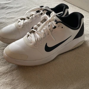 Vita Nike golfskor sneakers med svart swoosh - Snygga vita Nike sneakers med svart swoosh på sidan. Skorna har en klassisk design med snörning och en bekväm passform. Perfekta för en sportig look på golfbanan