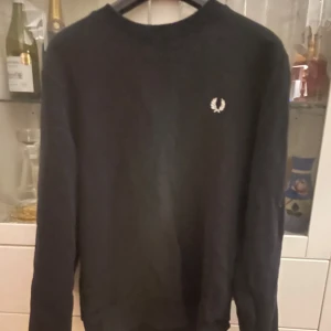 Svart tröja från Fred Perry - Säljer en stilren svart tröja från Fred Perry med deras ikoniska logga broderad på bröstet. Tröjan har långa ärmar och en rund halsringning. Perfekt för en klassisk och avslappnad look.