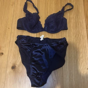Mörkblå spets-bh och trosor 70B samt 36 - Snygg mörkblå bh med spetsdetaljer och matchande trosor. Bh:n har justerbara axelband och en liten rosett mellan kuporna. Trosorna är i ett glansigt material som matchar bh:n perfekt. Perfekt set för en elegant look. I mycket fint skick, vintage och går inte att köpa i butik 