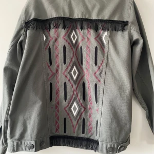 Militär grön  jeansjacka med mönster och fransar - Snygg grå jeansjacka från Unique You med coolt geometriskt mönster i rött, svart och vitt på ryggen. Jackan har fransdetaljer och knappar framtill. Super unik och coolt jacka! 