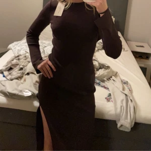 Mörkbrun klänning från NA-KD - Snygg mörkbrun klänning från NA-KD med lång ärm och en slit på vänster ben. Klänningen har en öppen rygg samt en söm vid bysten. Den går inte hela vägen ner på mig utan lite längre ner än mitten på smalbenen, jag är 169cm lång☺️