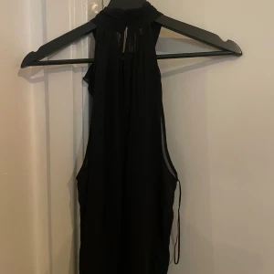 Svart topp med volangdetalj - Svart topp med volangdetalj vid halsen. Klänningen har en stilren design och är perfekt för festliga tillfällen. Den är ärmlös och har en smickrande passform.