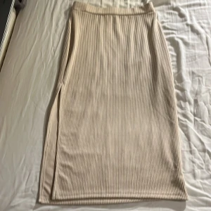 Beige ribbad långkjol - Snygg beige långkjol med ribbad textur. Kjolen har en stilren design med en diskret slits på sidan som ger extra rörelsefrihet. Perfekt för en elegant och avslappnad look.