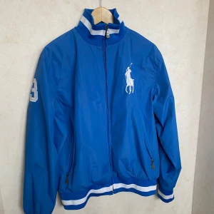 Blå jacka från Ralph Lauren - Snygg blå jacka från Ralph Lauren med dragkedja och ribbade muddar. Jackan har en broderad logga på bröstet och nummerdetalj på ärmen. Perfekt för en sportig look. Har dock ett hål under armen men det är inget som syns
