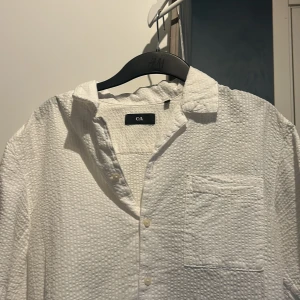Vit skjorta från C&A - Säljer en stilren vit skjorta från C&A med en texturerad yta. Skjortan har en bröstficka och knappar framtill. Perfekt för en avslappnad men ändå snygg look.