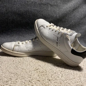 Vita Adidas Stan Smith sneakers - Klassiska vita Adidas Stan Smith sneakers med svart detalj på hälen. Skorna har snörning och är tillverkade i skinn. Perfekta för en stilren look. Otroligt bra nu till våren. Priset kan gå ner 