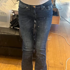 Mörkblå jeans från True Religion - Snygga mörkblå jeans från True Religion med ikoniska bakfickor och silverfärgade knappar. De har en tight passform och låg midja, perfekt för en trendig look. Jeansen är tillverkade i ett slitstarkt denimtyg. 