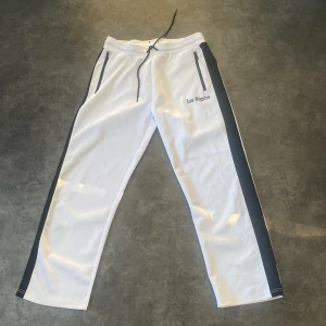 Vita träningsbyxor/ streetpants  med svarta detaljer - Snygga vita träningsbyxor/streetpants  med svarta sidopaneler och texten 'Los Angeles' broderad på framsidan. Byxorna har en elastisk midja med dragsko och praktiska dragkedjefickor.