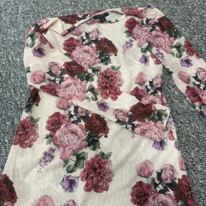 Blommig topp från Amisu - Säljer denna blommig topp från Amisu i storlek M. Toppen har ett färgglatt mönster med rosor i rosa, rött och lila på en ljus bakgrund. Den har långa ärmar och en elegant passform. Perfekt för att lysa upp garderoben! Genomskinlig vilket gör den lite mer festlig.