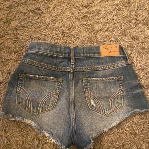 Jeans shorts från Hollister - Snygga blå jeansshorts från Hollister med slitna detaljer och fransig kant. Perfekta för en avslappnad sommarlook. Klassisk femficksdesign med knapp och dragkedja framtill. Storlek 25W