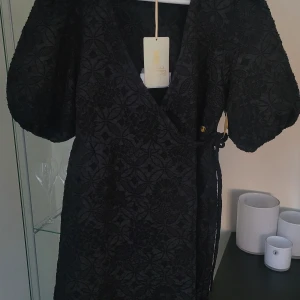 Svart omlottklänning med blommigt mönster - Elegant svart omlottklänning med ett vackert blommigt mönster. Klänningen har korta ärmar och ett knytband i midjan för en smickrande passform. Perfekt för en stilfull look.