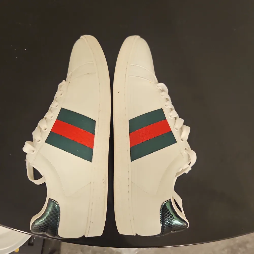 Snygga vita sneakers från Gucci med klassiska gröna och röda ränder på sidan. Skorna har snörning och en unik detalj i form av en mörkblå, ormskinnsmönstrad häl. Perfekta för en stilren look.. Kengät.