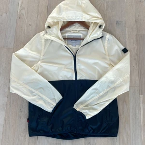 Beige och svart vindjacka från Woolrich - Säljer en snygg beige och svart vindjacka från Woolrich. Jackan har en praktisk huva och dragkedja framtill. Perfekt för blåsiga dagar och har en cool detalj med en fyrkantig logotyp på ärmen.