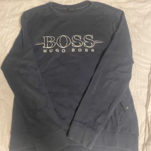 Mörkblå tröja från Hugo Boss - Säljer en stilren mörkblå tröja från Hugo Boss med deras ikoniska logga tryckt på framsidan. Tröjan har långa ärmar och en rund halsringning. Perfekt för en avslappnad och trendig look.