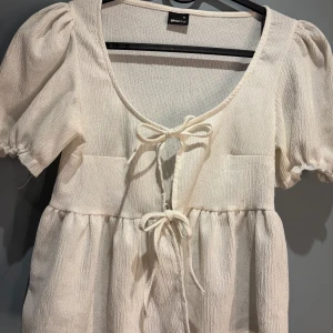 Vit blus från Gina Tricot - Säljer en söt vit blus från Gina Tricot i storlek XS. Blusen har korta puffärmar och knytband framtill som ger en romantisk touch. Materialet är lätt och luftigt, perfekt för varmare dagar.