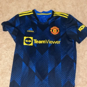 Blå fotbollströja från Adidas - Säljer en blå och svart fotbollströja från Adidas med korta ärmar. Tröjan har gula detaljer och ett mönster i blått och svart. Den har trycket 'Ronaldo 7' på ryggen och klubbmärket på bröstet. Perfekt för fotbollsfans!