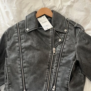 Helt ny, oanvänd med prislapp! Grå skinnjacka från Zara - Snygg grå skinnjacka från Zara med coola dragkedjedetaljer och knappar. Jackan har en croppad passform och långa ärmar. Perfekt för en edgy look!