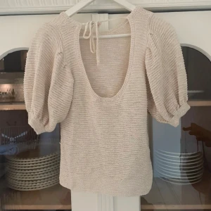 Beige stickad topp med puffärmar - Säljer en stilren beige stickad topp med puffiga trekvartsärmar och rund halsringning. Toppen har en knytning i nacken som ger en extra detalj. Perfekt för en chic och avslappnad look. Toppen har tyvärr inte kommit till användning och är därför i mycket bra skick