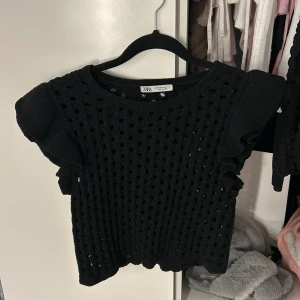 Svart stickad topp från Zara - Snygg svart stickad topp från Zara med hålmönster och volangdetaljer på ärmarna. Perfekt för att ge en trendig touch till din outfit. Toppen har en rund halsringning och korta ärmar.