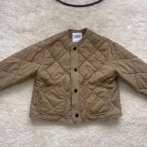 Beige quiltad jacka från Zara - Säljer en snygg beige quiltad jacka från Zara. Jackan har en rund halsringning och stängs med svarta knappar framtill. Den har två stora fickor på framsidan och är perfekt för kyligare dagar. Säljer då den inte satt som jag ville!💕💕