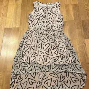 Ärmlös klänning med hjärtmönster - Söt ärmlös klänning med ett lekfullt hjärtmönster i svart på en ljus bakgrund. Klänningen har en luftig passform och är perfekt för varma dagar. Den har en elastisk midja som ger en fin silhuett.