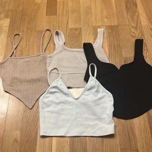 Fyra olika toppar i olika färger - Säljer fyra snygga toppar i olika stilar och färger. En beige ribbad topp med smala axelband, en vit v-ringad topp, en svart topp med breda axelband och en beige topp med breda axelband. Perfekta för sommaren!