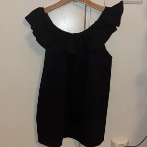 Svart offshoulder topp med volang - Snygg svart offshoulder topp med volangdetalj runt halsen. Perfekt för en stilren look. Toppen är ärmlös och har en luftig passform.