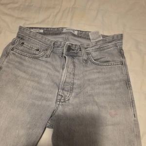 Grå jeans från Jack & Jones - Säljer ett par grå jeans från Jack & Jones i loose fit. De har en klassisk femficksdesign och knappgylf. Perfekta för en avslappnad stil. Obs färgfläckar fram och lite bak annars i bra skick