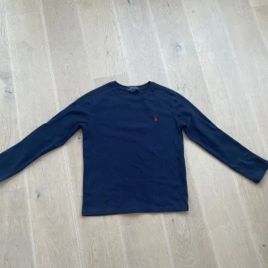 Mörkblå långärmad tröja från Polo Ralph Lauren - Säljer en stilren mörkblå långärmad tröja från Polo Ralph Lauren med det klassiska röda broderade logotypen på bröstet. Perfekt för en avslappnad men ändå elegant look. Storlek M i barn storlek