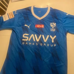 Blå fotbollströja från Al Hilal / Neymar - Snygg blå fotbollströja från Puma med Al Hilal-logga och sponsortryck. Tröjan har korta ärmar och ett stilrent mönster. Perfekt för fotbollsfans som vill visa sitt stöd. Tröjan har Neymar Jr nr 10 på ryggen