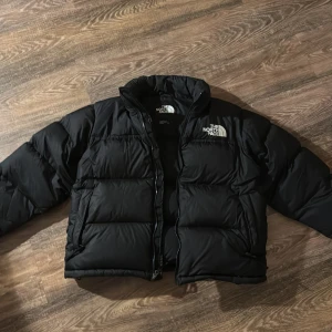 Svart dunjacka från The North Face - Säljer en svart dunjacka från The North Face. Jackan har en pufferdesign med dragkedja framtill. Den är långärmad och har en klassisk logga på bröstet. Köpt för 2 år sen, knappt använd. Nypris 3500kr, säljer för 1000kr. 